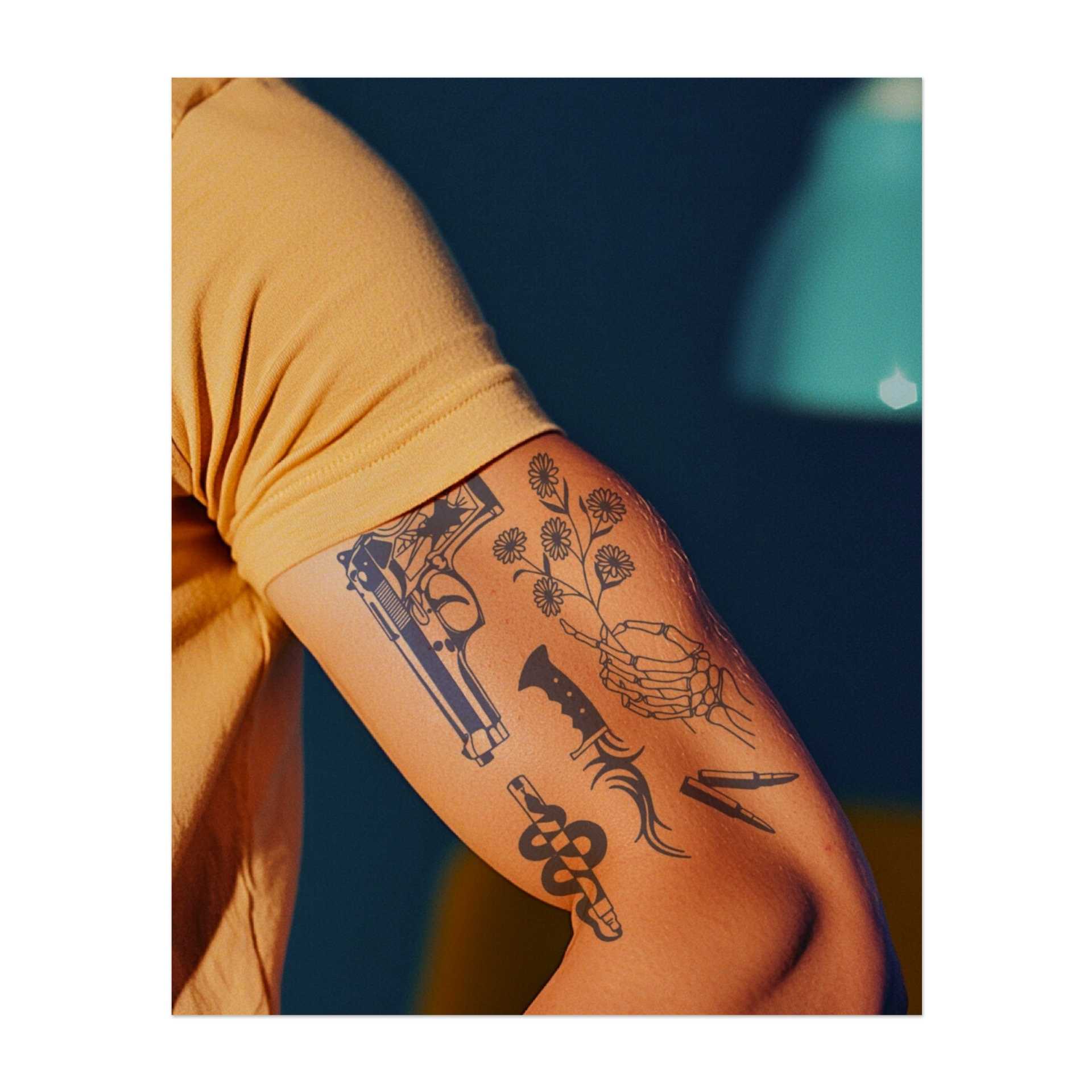 Ein Bündel kontrastreicher Symbole: eine Pistole, ein Messer, eine Skeletthand, eine Schlange, ein Blumenstrauß und eine Kugel. Diese Designs repräsentieren das Gleichgewicht zwischen Leben und Tod, Gefahr und Schönheit und fangen sowohl Eleganz als auch rohe Kraft ein.