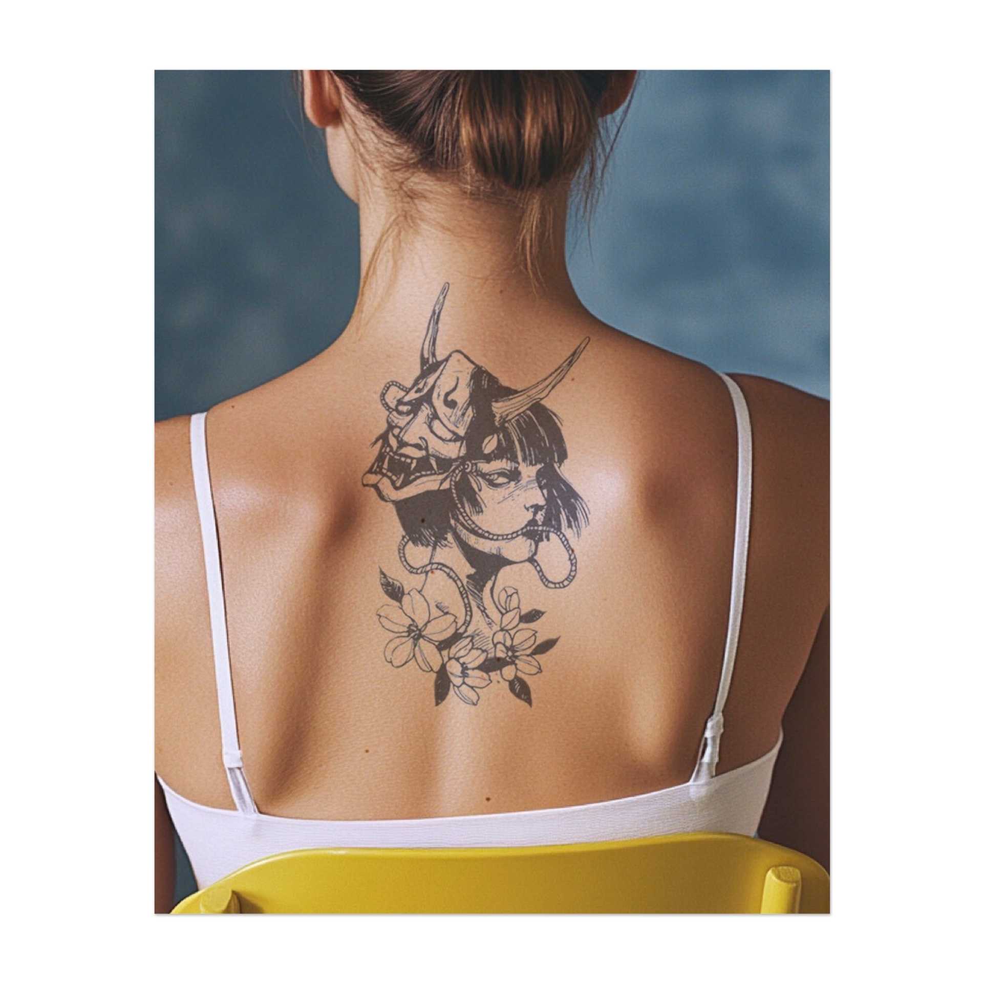 Ein auffälliges Tattoo einer Frau, die eine traditionelle Oni-Maske trägt, umgeben von zarten Blumen. Das Design kombiniert Schönheit und Dunkelheit und symbolisiert Stärke, Geheimnis und Transformation. Perfekt für jemanden, der sowohl Macht als auch Anmut umarmt.