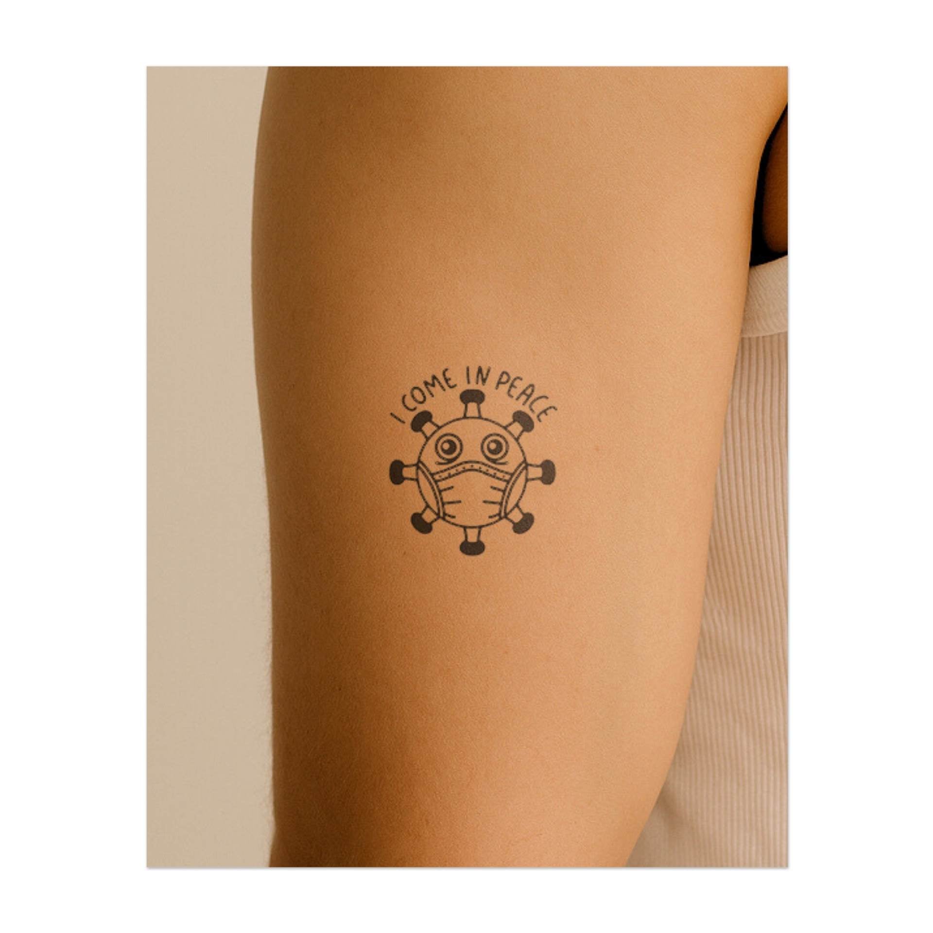 Un virus de dessin animé ludique avec un masque facial sur lequel est écrit « JE VIENS EN PAIX ». Ce tatouage apporte de l'humour à cette époque et symbolise la résilience et les bonnes vibrations dans les moments difficiles.